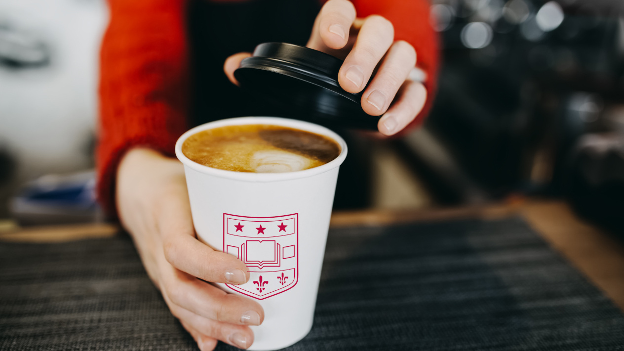 Barista in apron is holding in hands hot cappuccino in white takeaway paper cup. Coffee take away at cafe shop