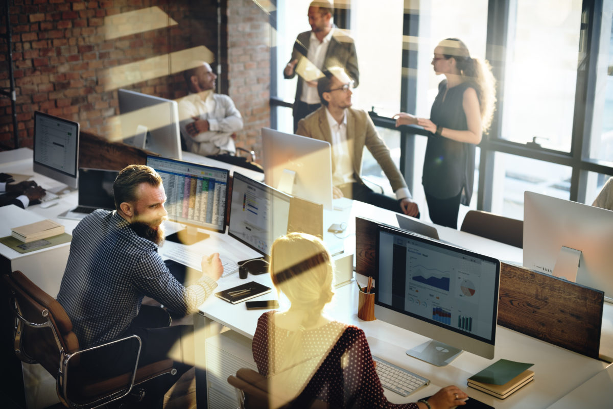 A group of office workers sitting at desks while colleagues stand around them.