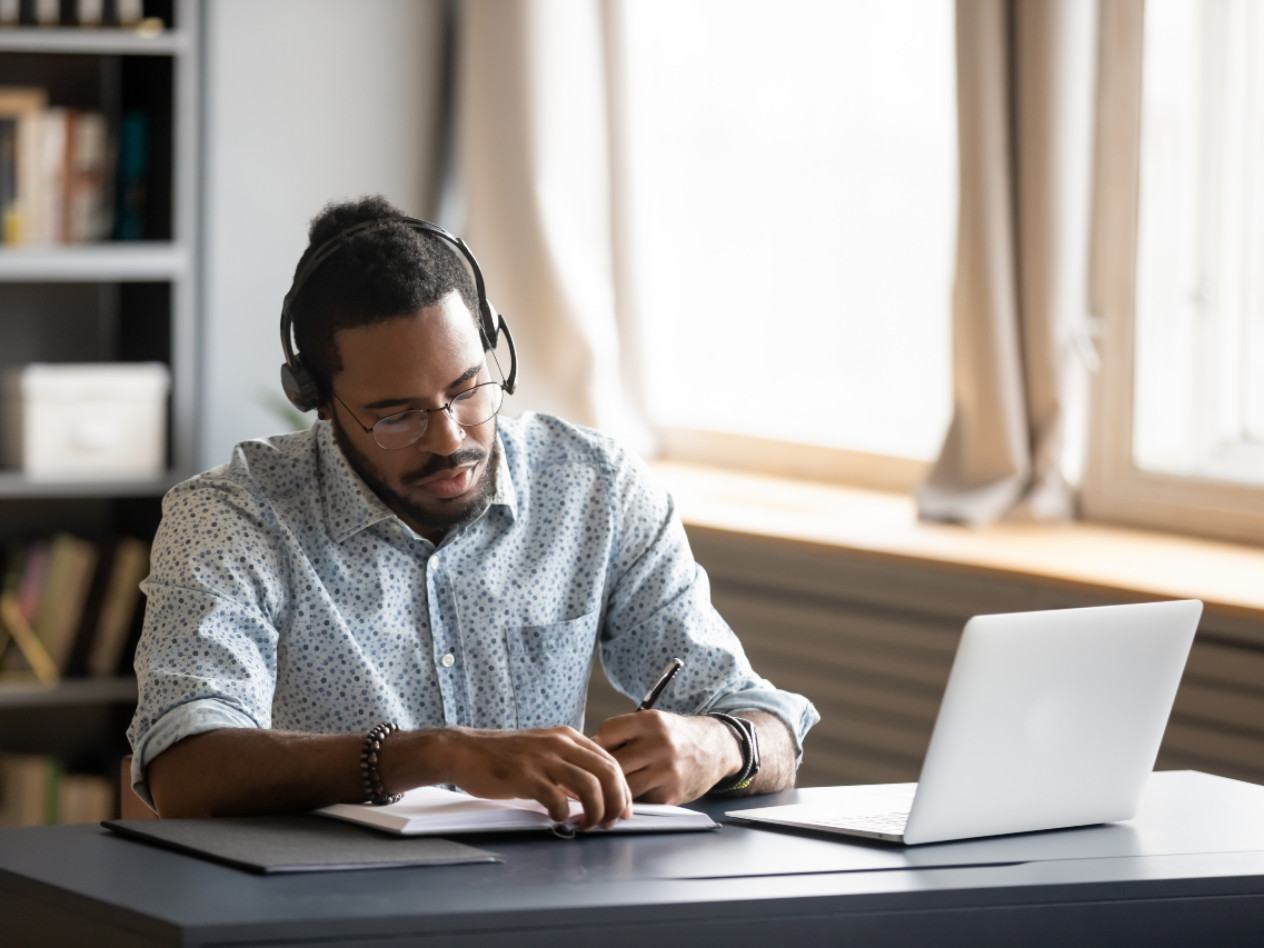A man wearing headphones sits at a desk in his home with an open computer in front of him. 