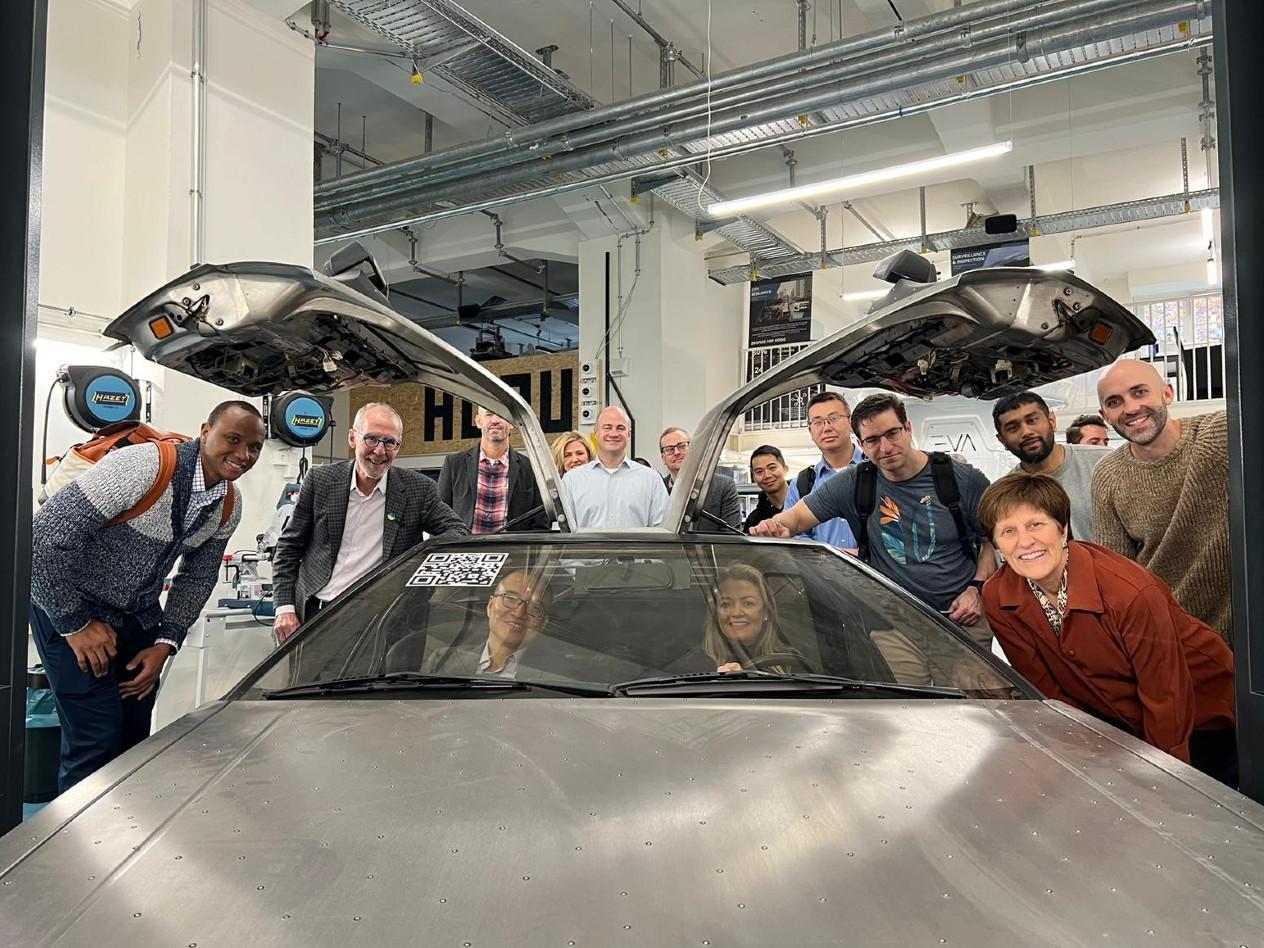 Students sit in a car with gull wing doors while others stand around it