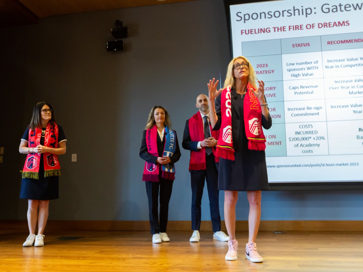 Four EMBA students with red scarves standing on stage in front of a presentation screen.