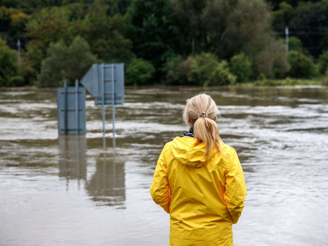 Woman wearing yellow raincoat looks at floodwater covering road.