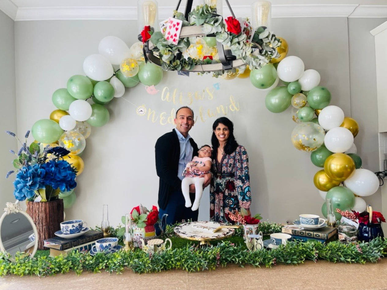 Parents hold a child as they stand at a table with decorations and balloons