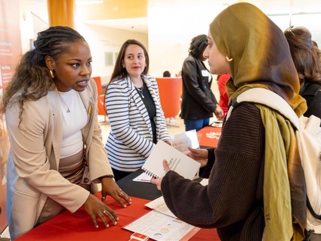 A woman stands at a table speaking to another woman.