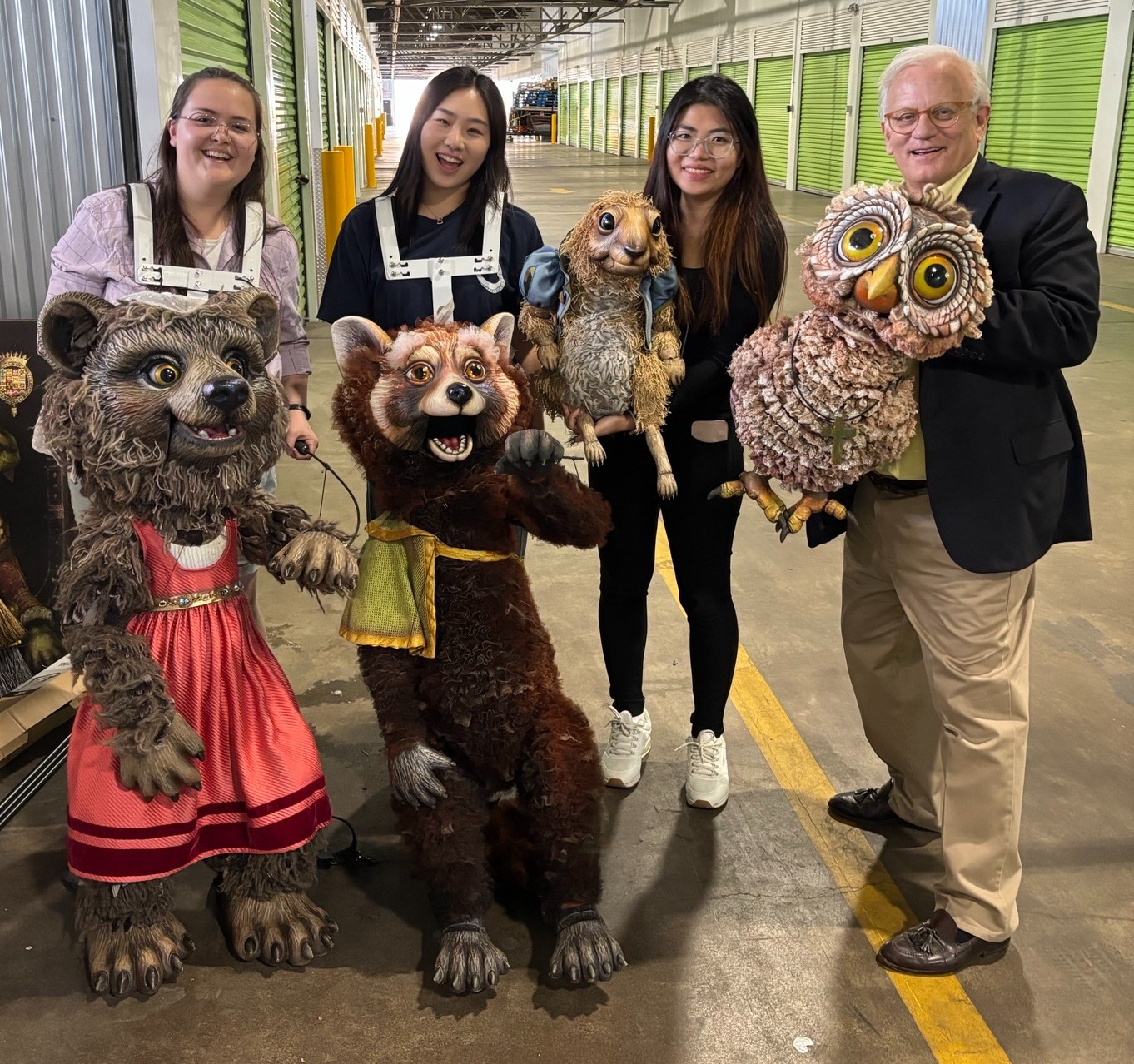 Four people standing in a warehouse, holding large animal puppets.